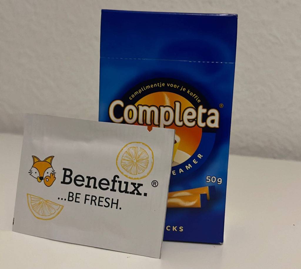 Completa Kaffeeweißer im Test steht in seiner blauen Verpackung hinter verpacktem Benefux Be Fresh Erfrischungstuch vor weißer Fläche.
