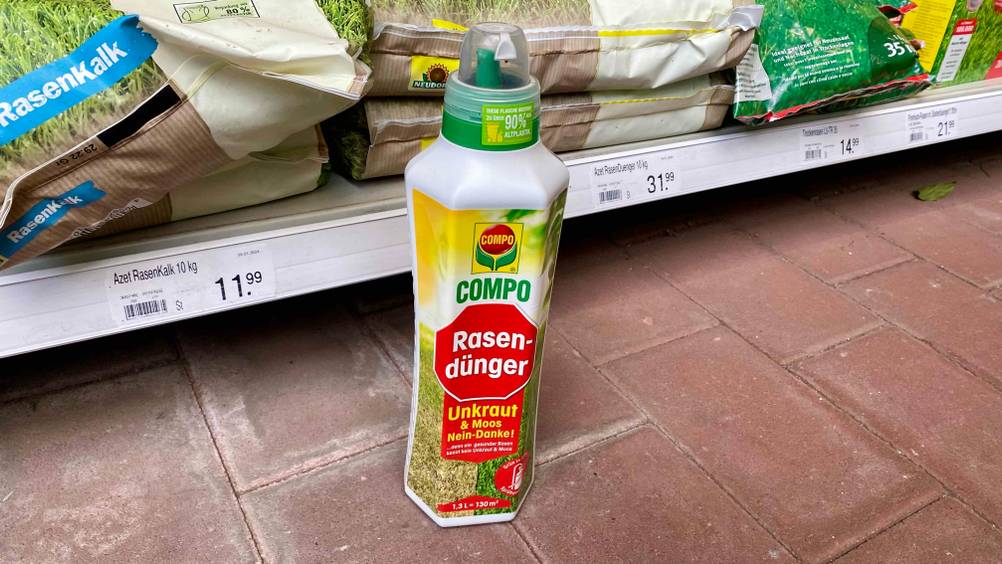Im Test: Eine Flasche Rasendünger gegen Unkraut steht vor einem Verkaufsregal mit Preisschildern auf einem Steinboden.