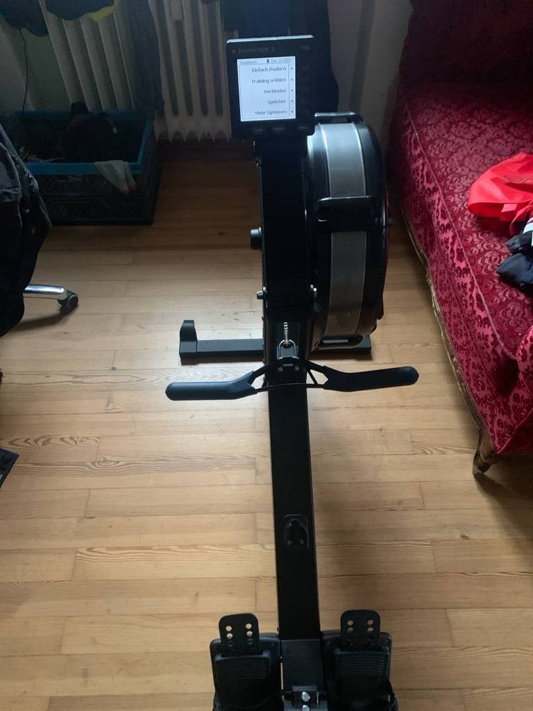Ansicht von oben auf ein schwarzes Indoor-Rudergerät im Test mit eingeschalteter LCD-Anzeige in einem Wohnbereich auf Parkett neben einem roten Sofa.