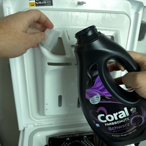 Aus der Verschlusskappe des Waschmittels für schwarze Wäsche von Coral wird Waschmittel in das Waschmittelfach geschüttet.