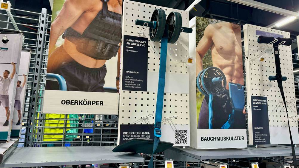 Bauchtrainer im Test: Ein Gerät hängt zwischen Infoplakaten an einer Lochwand.