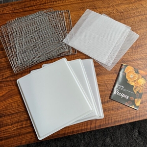 Die Gitter sowie einige Einlagen des Cosori CP267-FD liegen neben dem Rezeptbuch auf einem Tisch