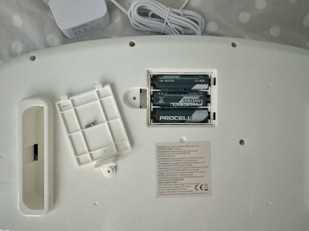 Kinderkeyboard-Test: Detailansicht der weißen Unterseite mit Fokus auf das Batteriefach inklusive 3 AA-Batterien und ein danebenliegendes weißes Adapterkabel.