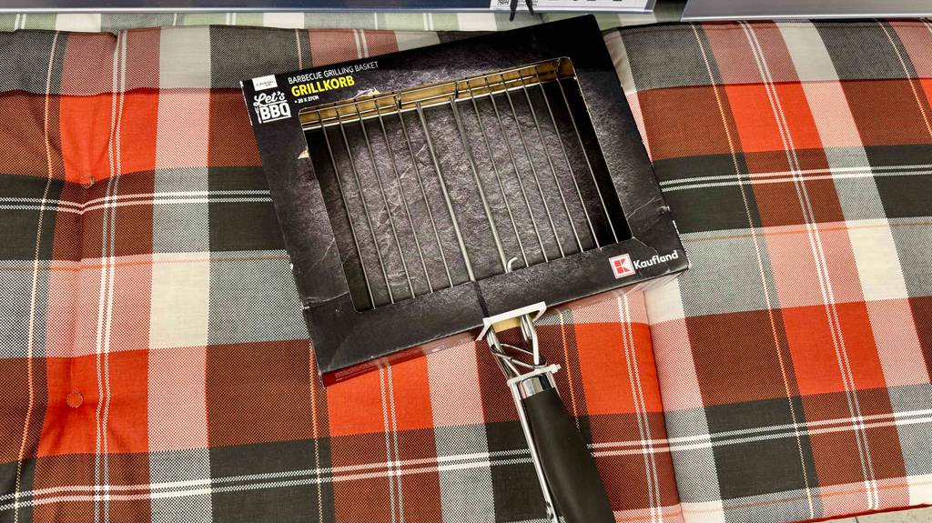 Getestete: Ein Grillkorb mit schwarzem Kunststoffgriff liegt in seiner Verpackung auf einem karierten Stuhlkissen.