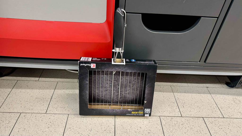 Vor einem Regal steht eine getesteter Grillkorb mit schwarzem Kunststoffgriff in seiner Verpackung.