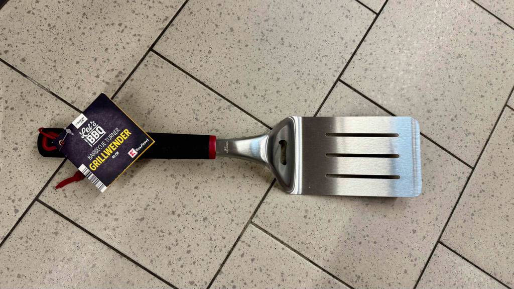 Ein getesteter Grillwender mit schwarzem Kunststoffgriff inklusive Öse zum Aufhängen liegt auf dem Boden.