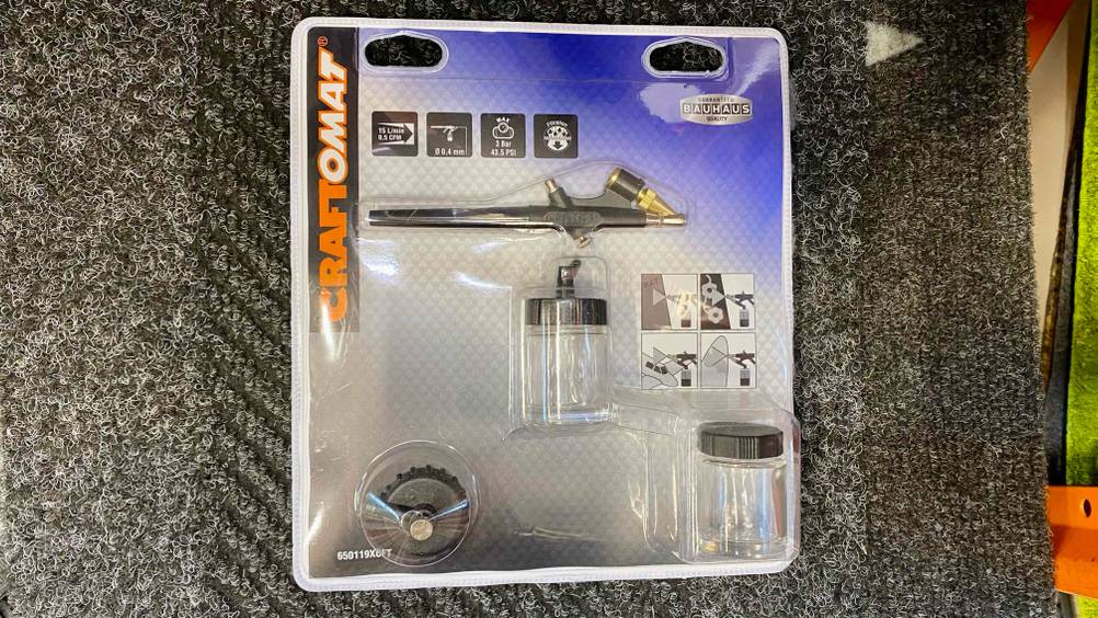 Eine verpackte Airbrushpistole im Test liegt auf einem grauen Teppich.
