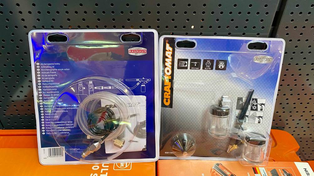Im Test: Eine Airbrushpistole und ihre Zubehörschläuche beide in Originalverpackung an einem grauen Lochbrett lehnend.