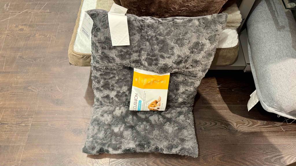 An einem Hundebett auf dem Regal lehnt ein getestetes Hundekissen aus grauem, flauschigen Material.