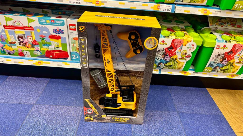 Vor einem Regal mit Bau-Spielzeug steht der gelbe getestete Spielzeug-Kran mit Fernbedienung innerhalb seiner Verpackung inklusive Sichtfenster.