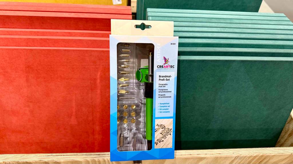 Das Brandmalerei-Set im Test steht innerhalb der Verpackung vor roten und grünen Spanplatten im Baumarkt.