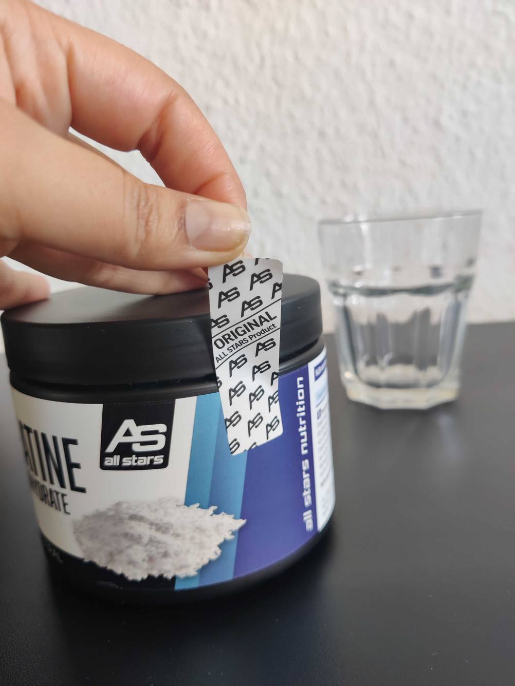 Eine Hand entfernt das Sicherheitssiegel an der Dose des getesteten Creatin-Pulvers, im Hintergrund ein Glas.