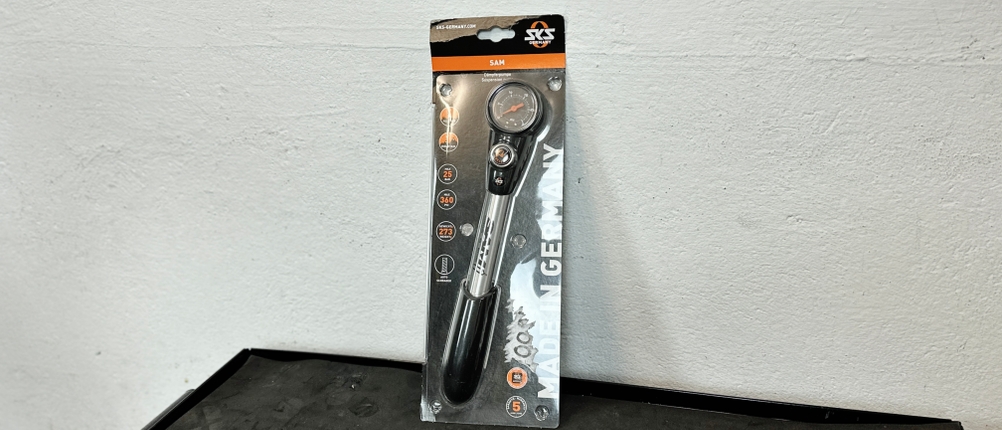 Die SKS-SAM-Dämpferpumpe ist originalverpackt und steht auf einer Werkbank. Das Produkt ist im Blister sichtbar, mit Bedruckung und Logo.