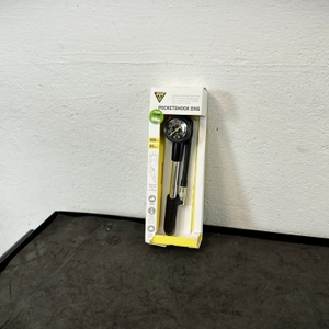 Die Topeak Pocketshock DXG steht originalverpackt auf einer Werkbank. Die Verpackung ist weiß-gelb mit Sichtfenster.