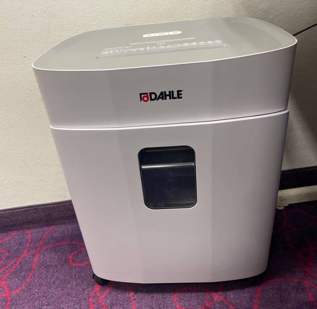 Ansicht von vorn auf einen weißen getesteten Aktenvernichter inklusive Sichtfenster, der Marke Dahle, welcher auf einem violettfarbenen Teppich vor weißer Wand steht.