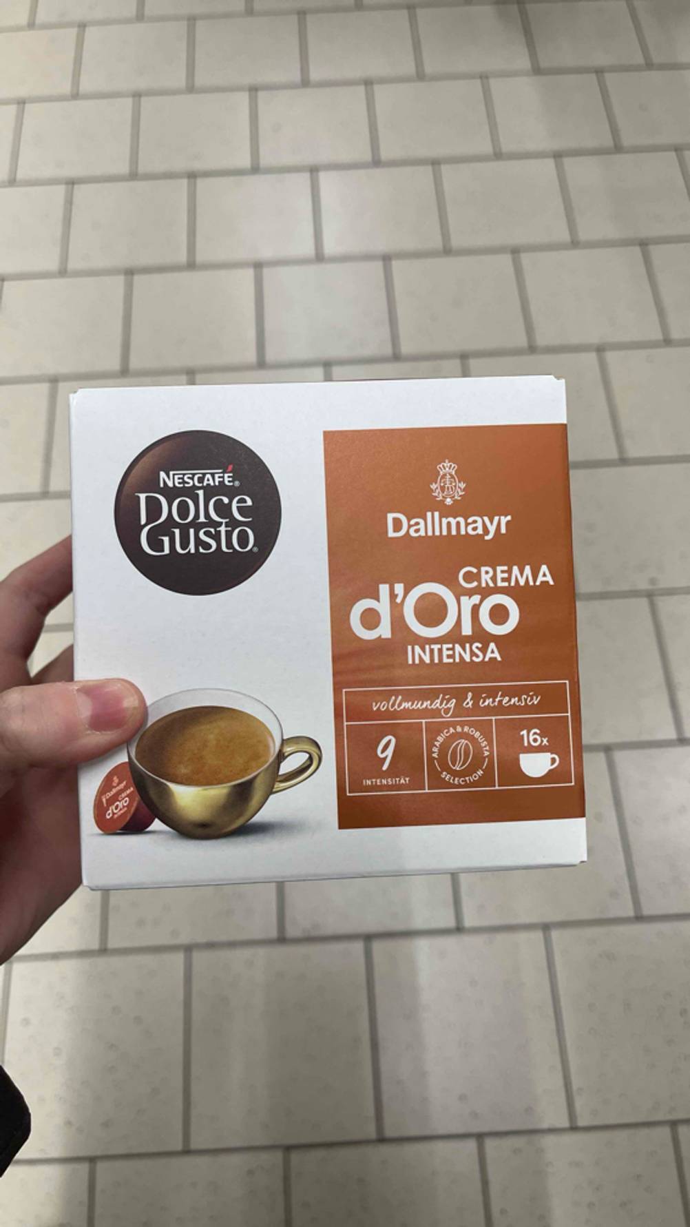 Getestet: Die Dallmayr-Kapseln der Geschmacksrichtung Crema d’Oro Intensa befinden sich in einem weiß-braunen Karton, der in einer Hand über einem weißen Fliesenboden gehalten wird.