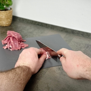Ein rohes Stück Fleisch liegt auf einem Schneidebrett, das Fanteck Gyuto wird gerade angesetzt. Die linke Hand fixiert das Fleisch für den ersten Schnitt.