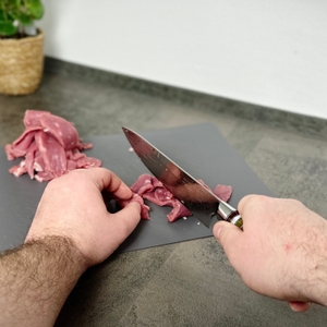 Zwei Hände schneiden rohes Fleisch mit dem Fanteck Gyuto auf einem Kunststoff-Schneidbrett. Der Fokus liegt auf dem Schneidevorgang mit sichtbarer Klingenspitze.