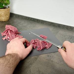 Eine Hand führt das Fanteck Gyuto beim Schneiden von rohem Fleisch auf einem dunklen Schneidbrett. Die zweite Hand richtet die Schnittführung aus.