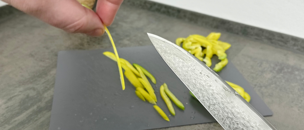 Eine gelbe Paprika wird auf einem grauen Schneidbrett in feine Streifen geschnitten. Das Fanteck Gyuto liegt im Vordergrund, die Spitze zeigt zur Kamera.