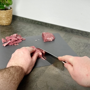 Das Santoku-Messer wird angesetzt, um ein Stück Fleisch zu schneiden. Eine Hand hält das Fleisch, die andere führt das Messer.