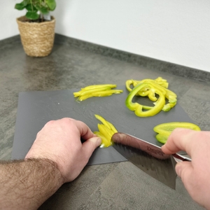 Mit dem Schneidwerk-Santoku-Messer werden gelbe Paprikastreifen auf einem grauen Brett vorbereitet. Die linke Hand richtet das Gemüse aus.