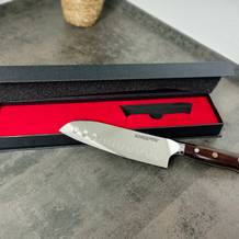 Das Schneidwerk Santoku-Messer liegt auf einer schwarzen Produktverpackung mit rotem Inlay. Klinge und Griff sind vollständig zu sehen.