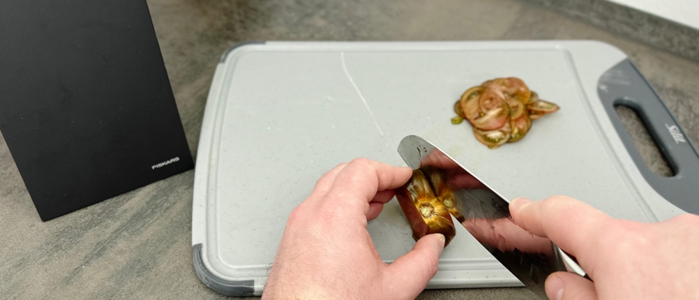 Das Schneidwerk-Santoku-Messer schneidet eine dunkle Tomate auf einem Kunststoffbrett. Die Klinge liegt eng an der Schnittfläche.