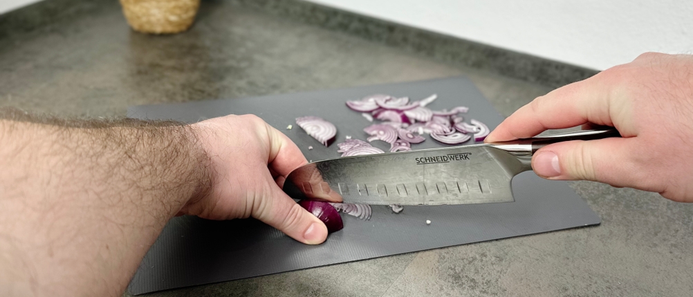 Das Santoku-Messer von Schneidwerk schneidet eine rote Zwiebel in feine Streifen. Die Klinge liegt flach auf dem Brett.