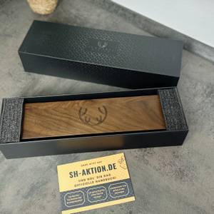 Die geschlossene Produktverpackung des Schwartzhirsch Caliburn zeigt die schwarze Außenbox mit eingelegter Holztruhe. Daneben liegt eine gelbe Infokarte mit QR-Code.