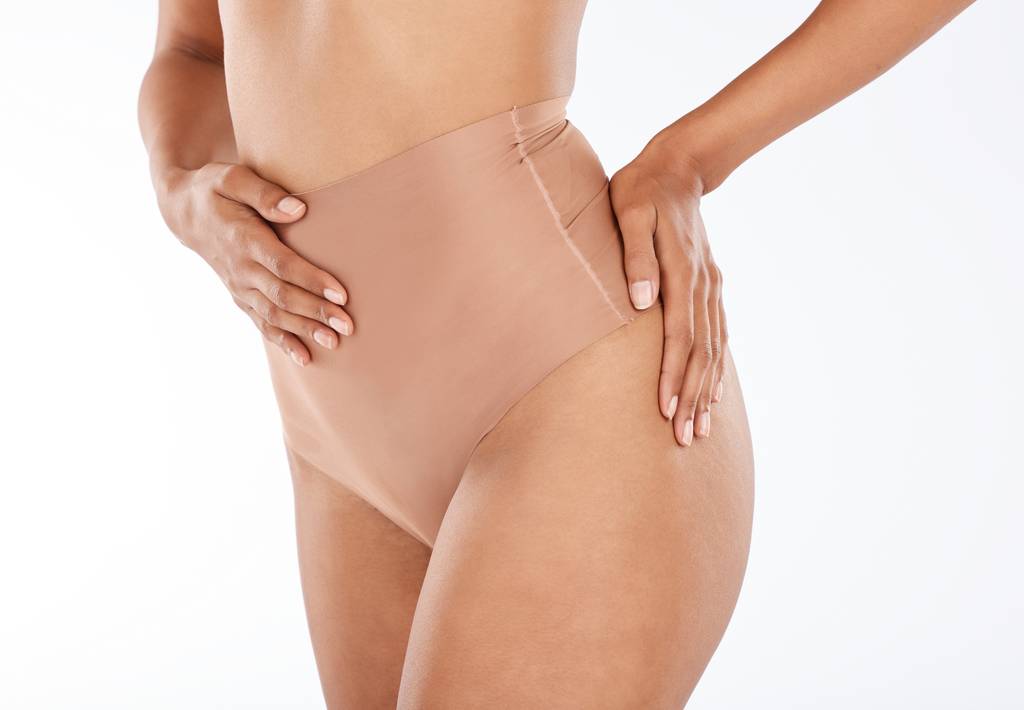 Damen-Shapewear-Test: Eine Frau präsentiert Shapewear vor einem weißen Studiohintergrund, wobei der Fokus auf der Bauchpartie und der ästhetischen Darstellung von Gewichtsverlust liegt. 