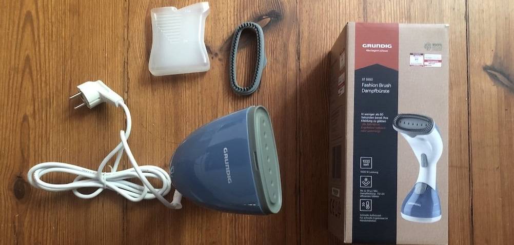 Die Verpackung und das Zubehör des Grundig Fashion Brush Dampfglätters liegen nebeneinander auf dem Boden.