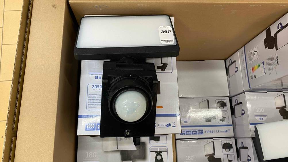 Ein rechteckigen Darius LED-Strahler im Test, mit Fokus auf den integrierten Bewegungsmelder liegt, mit einem Preisaufkleber versehen, auf Verpackungen seinesgleichen in einem großen braunen Karton.