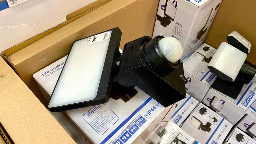 Ansicht auf einen rechteckigen Darius LED-Strahler im Test, inklusive Bewegungsmelder und Preisschild, auf Verpackungen des gleichen Modells in einem großen Lieferkarton.
