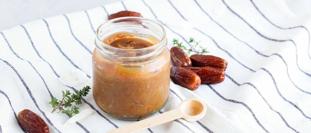 Ein Glas mit Dattelpaste steht auf einem Geschirrtuch mti Datteln und einem Holzlöffel daneben