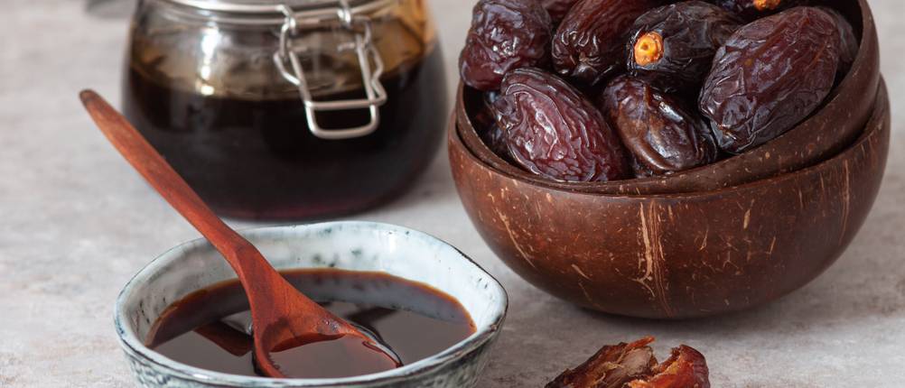 Eine Schale mit Dattelsirup und Löffel steht vor einem Einmachglas mit Dattelsirup und einer Holzschale mit Datteln