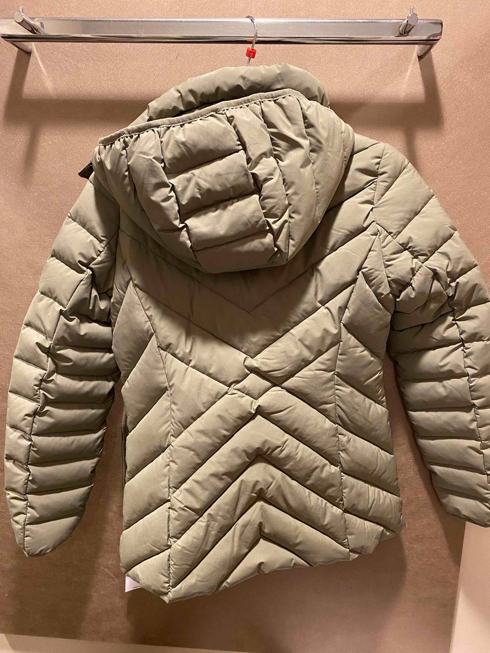 Daunenjacke für Damen in Beige mit Kapuze hängt mit der Rückseite nach vorne an einer Garderobe.