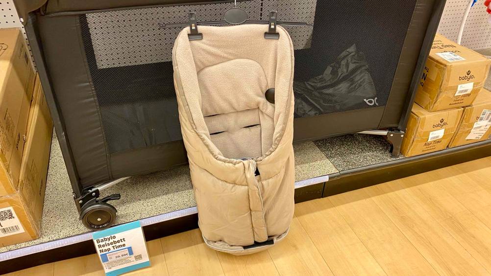 Ein getesteter Daunenschlafsack-Baby hängt an einem Bügel vor einem Babybett in einer Verkaufsfläche.