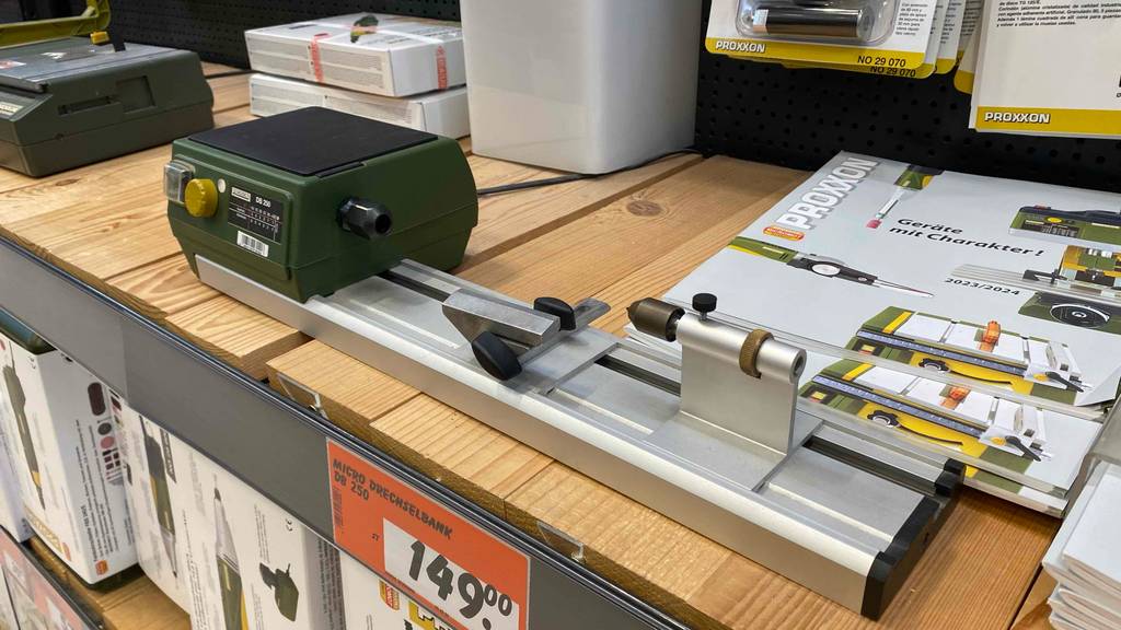 Gesamtansicht von schräg oben auf eine Proxxon Micro-Drechselbank im Test, mit grünem Motor-Gehäuse sowie Werkstück aus Aluminium und Preisschild in einem Ausstellungsregal.