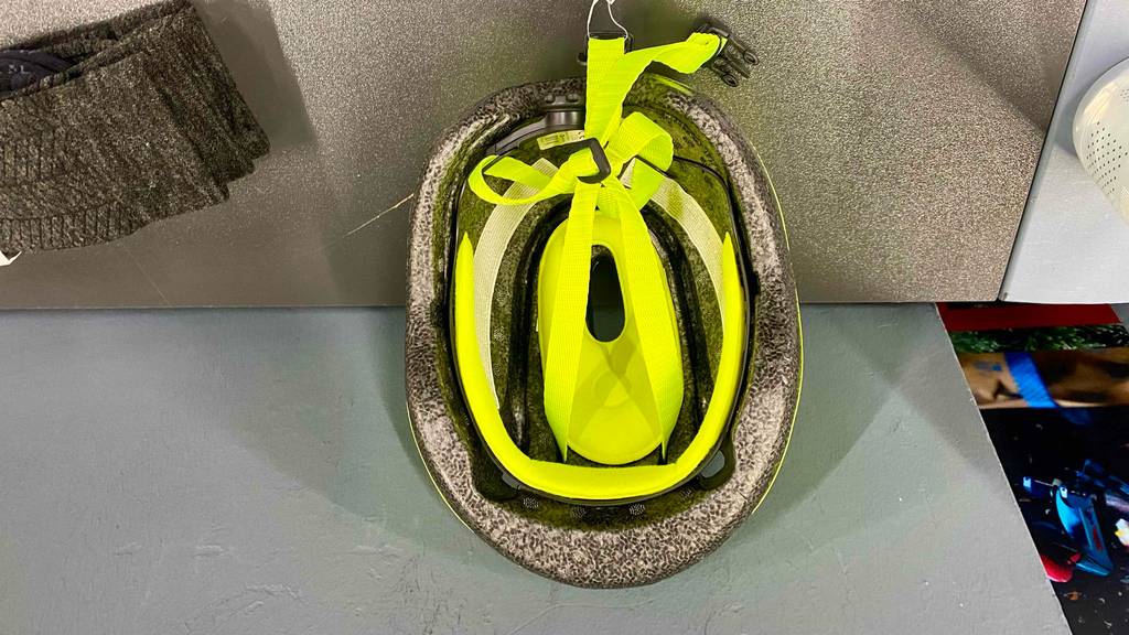 Ein hellgrau gesprenkelter, getesteter Baby-Fahrradhelm mit Fokus auf sein Inneres. Inklusive leuchtend gelber Befestigungsgurte und liegt auf einer grauen Oberfläche.