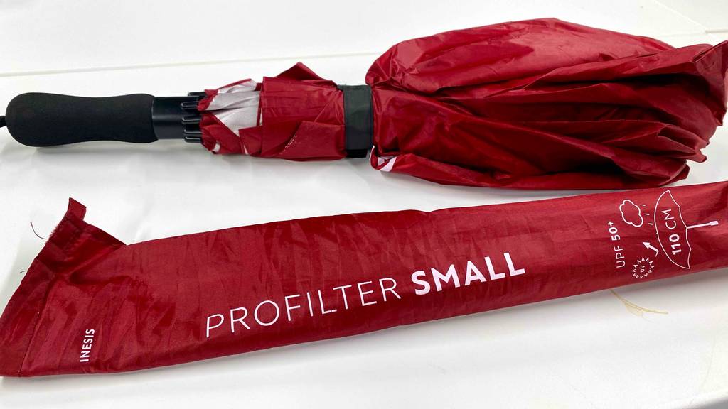 Golfschirm-Test: Ein roter Golfschirm mit Lichtschutzfaktor 50+ liegt geschlossen neben seiner roten Hülle auf einer weißen Oberfläche.