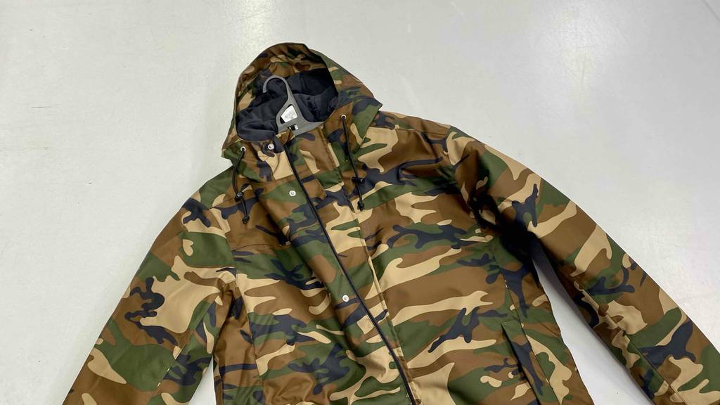 Eine getestete Herren-Jagdjacke im Camouflage-Muster liegt mit ausgebreiteten Ärmeln und Kapuze auf einer weißen Oberfläche.