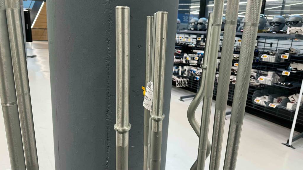 Mehrere getestete Langhantelstangen stehen aufgereiht vor einer Betonwand.