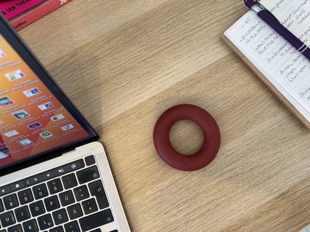 Ein Handtrainer-Ring liegt zwischen einem Laptop und einem Notizblock auf einem Holztisch.