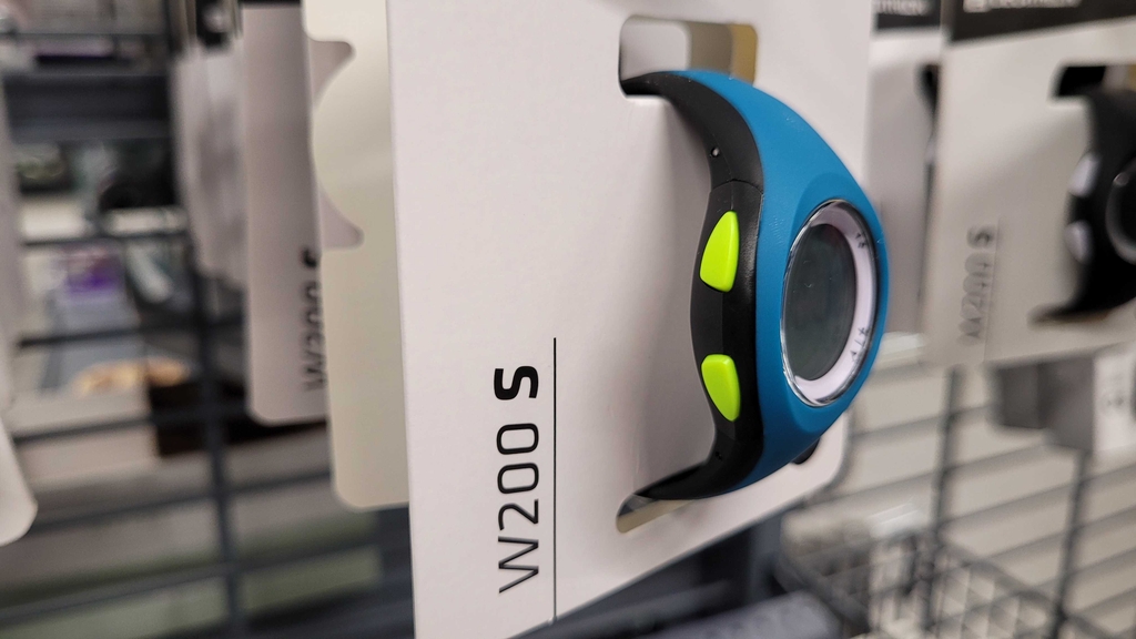 Seitliche Vorderansicht einer blau-schwarzen Sportuhr inklusive runder LCD-Anzeige und giftgrüner Einstelltasten in einem weißen Verpackungskarton im Verkauf.