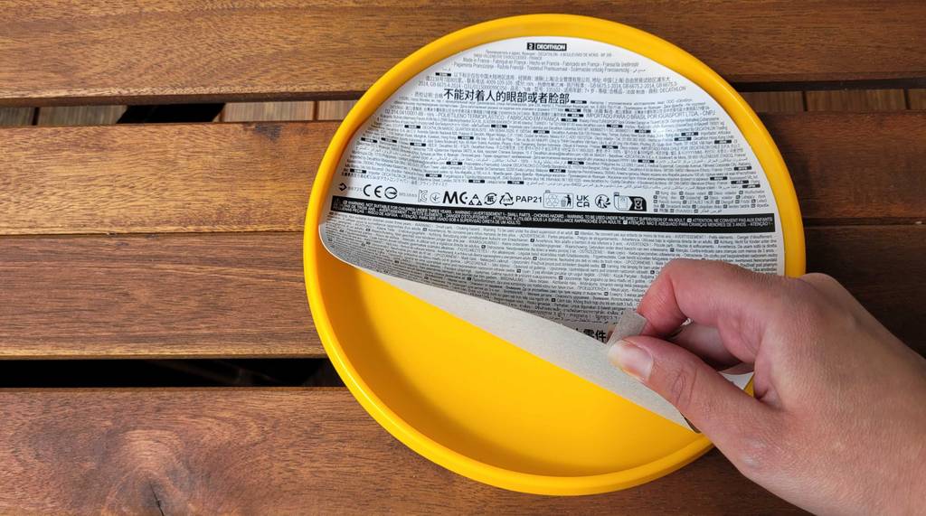 Eine Hand zieht den Verpackungskarton von einer gelben Frisbee-Scheibe, die auf einem Holzuntergrund liegt, ab.