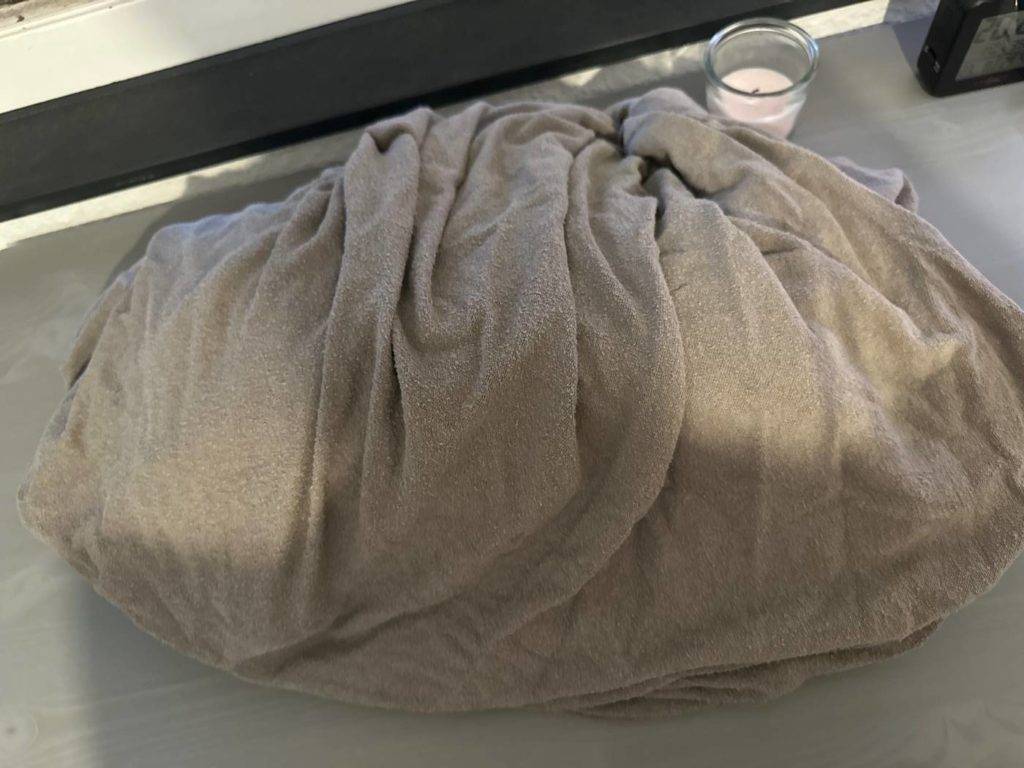 Ein getestetes Spannbettlaken in Grau aus Frottee liegt zusammengerollt auf einem Bett.