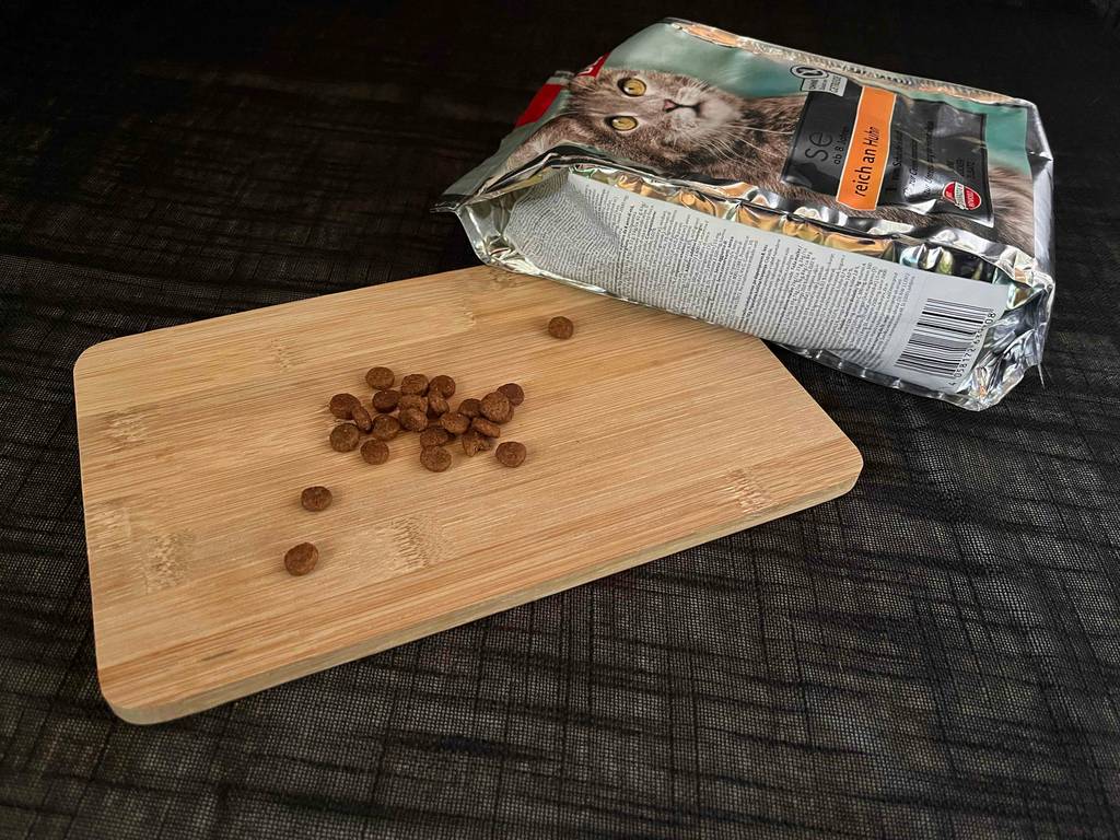 Getestet: Eine Handvoll Katzen-Trockenfutter liegt auf einem Schneidebrett, die Verpackung liegt quer im Hintergrund.
