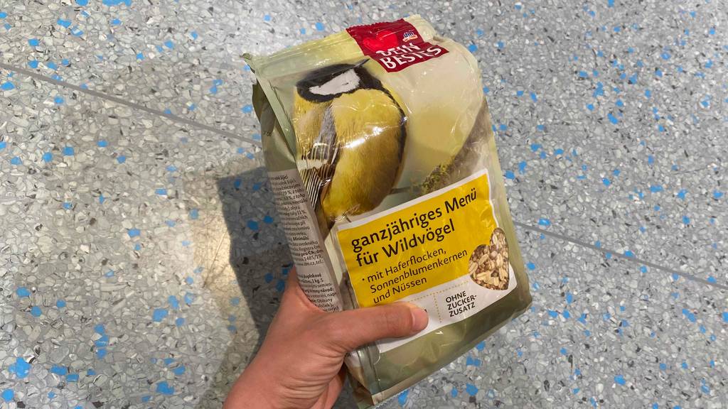 Getestet: Hand hält einen Beutel Dein Bestes Vogelfutter für Wildvögel mit Haferflocken und Nüssen über einem Fliesenboden.