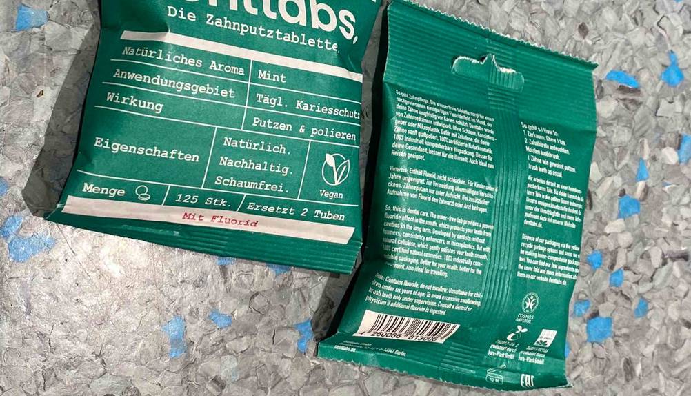 Zahnputztabletten im Test: zwei grüne Verpackungen liegen nebeneinander auf einer blau-grau gesprenkelten Oberfläche, eine mit der rückseitigen Produktbeschreibung nach oben.